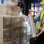A man in a yellow hard hat and safety vest stretch wraps a pallet of cardboard boxes in a warehouse.