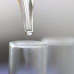 A close-up view shows a clear pipette dropping a clear liquid into a glass test tube. The background is blurry.
