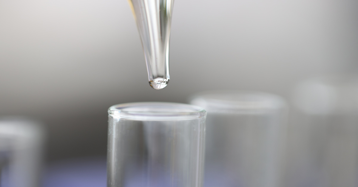 A close-up view shows a clear pipette dropping a clear liquid into a glass test tube. The background is blurry.
