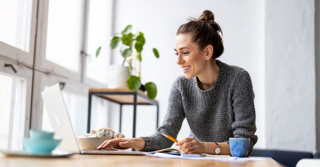 A young professional sitting in a nice living space and working on a sleek silver laptop while taking notes.