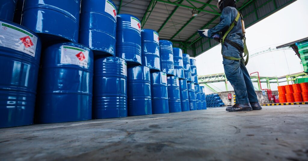 A person, who is wearing a blue jumpsuit and a yellow harness, is looking at a pile of metal barrels.