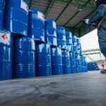 A person, who is wearing a blue jumpsuit and a yellow harness, is looking at a pile of metal barrels.
