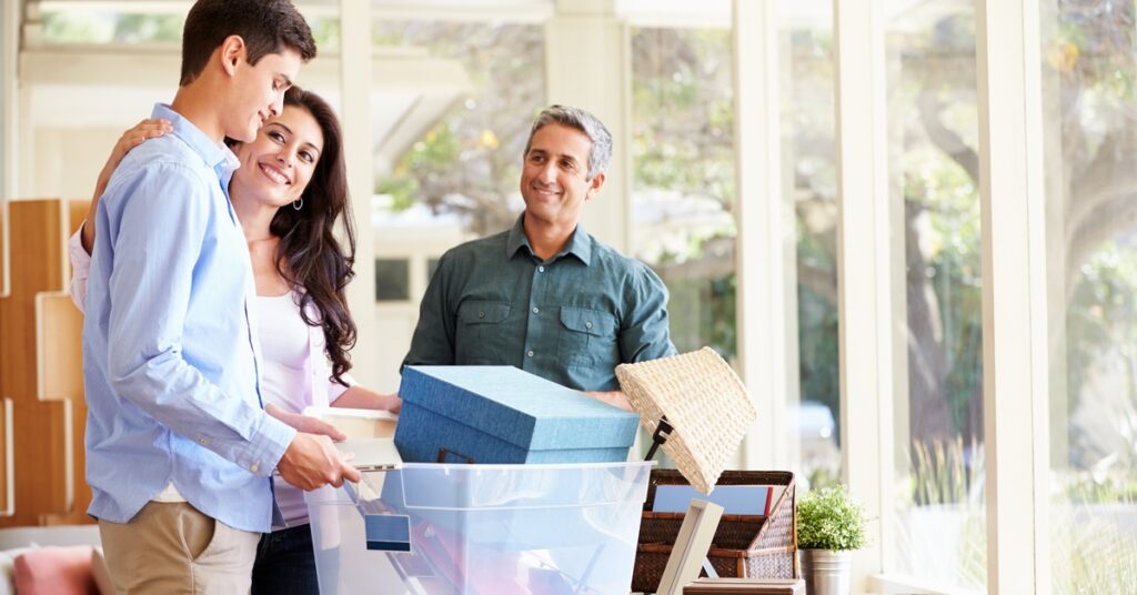 Parents helping their teenage son pack for college; both parents smile at their son as he packs his boxes.