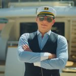A man wearing a sailor hat, a blue shirt, a blue vest, and aviator sunglasses standing on the back side of a small yacht.