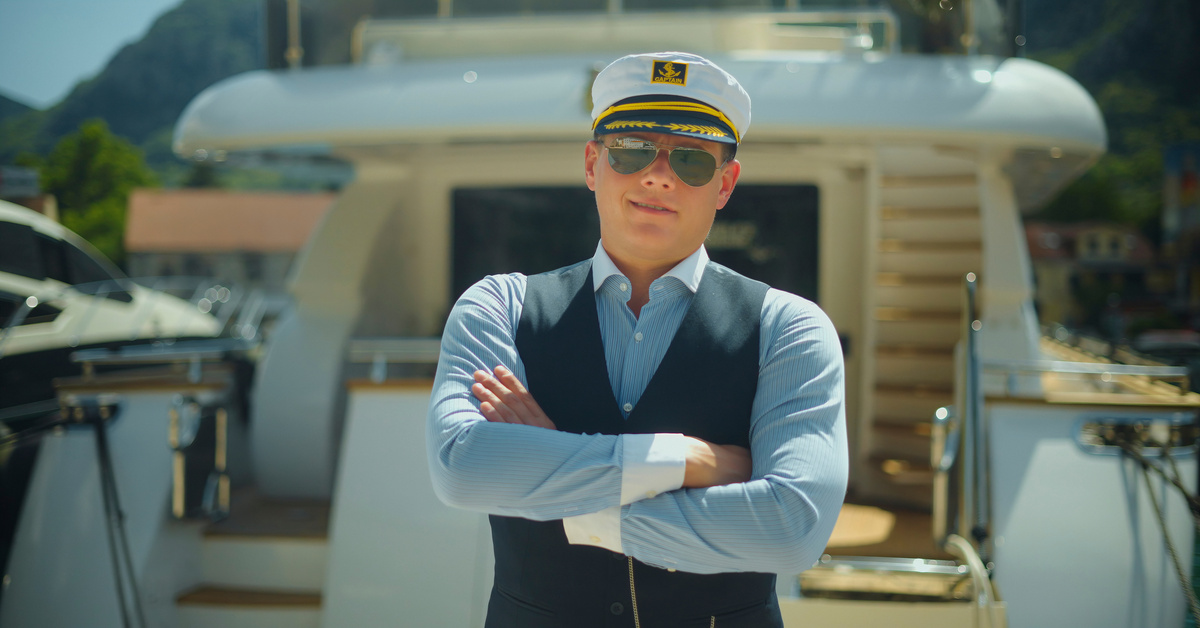 A man wearing a sailor hat, a blue shirt, a blue vest, and aviator sunglasses standing on the back side of a small yacht.
