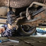 A person lying on their driveway during the daytime and using a wrench to make repairs underneath their car.