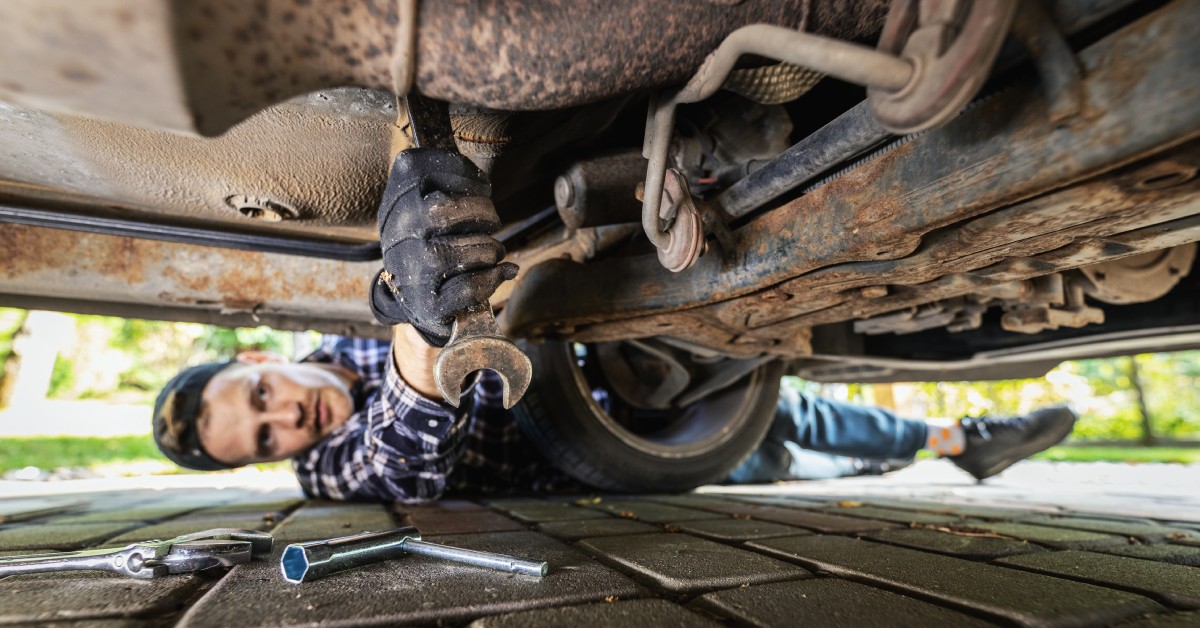 A person lying on their driveway during the daytime and using a wrench to make repairs underneath their car.