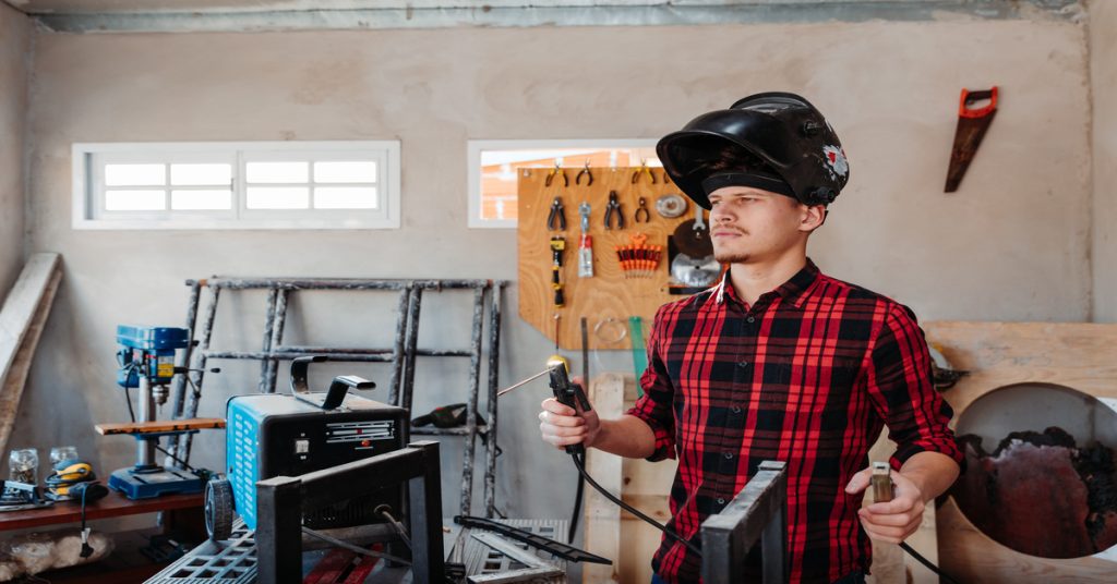 A young student welder wears a red and black plaid button-up and a welding helmet as they hold the welding gun.