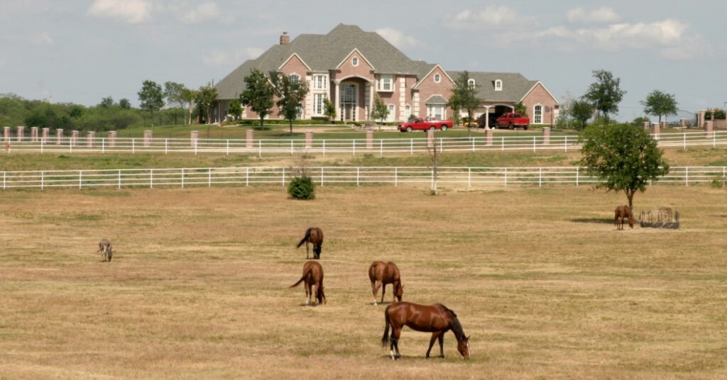 A large ranch house on a hill overlooks a fenced pasture where several horses graze under a pale blue sky.