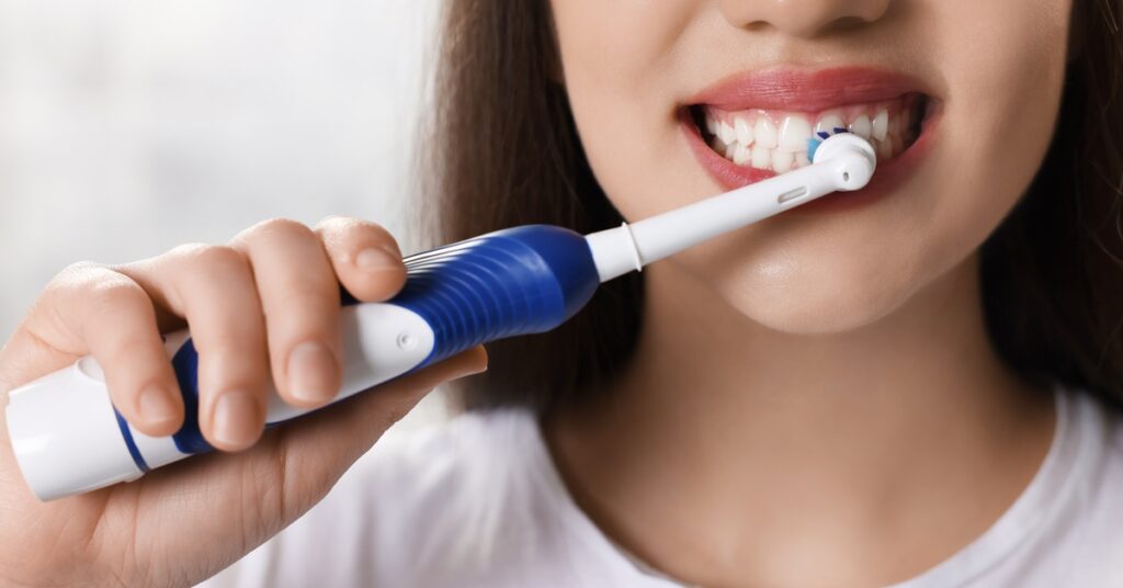 A close-up of a woman with brown hair brushing her teeth with an electric toothbrush. The background is white.