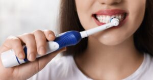 A close-up of a woman with brown hair brushing her teeth with an electric toothbrush. The background is white.