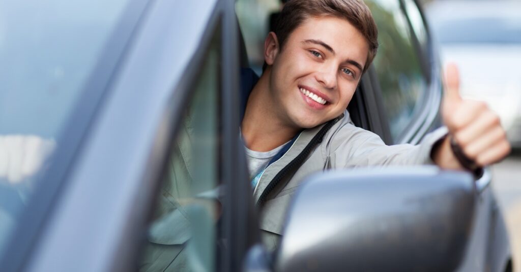 A young man sits behind the wheel of a car and leans out the window. He smiles and gives the thumbs-up gesture.