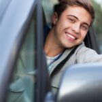 A young man sits behind the wheel of a car and leans out the window. He smiles and gives the thumbs-up gesture.