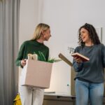 Two college-aged women unpack boxes and books in a bright dorm room, smiling and chatting as they move in together.