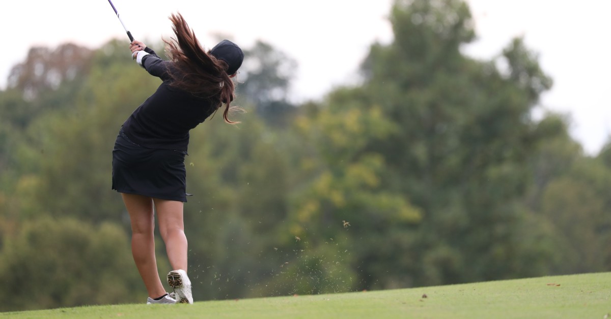 A high school student in a dark uniform swinging a golf club in a wide arch while standing on a manicured course.