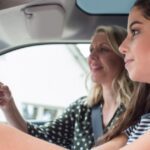 A teenager girl sits behind the wheel of a vehicle with her mother sitting beside her, pointing at the windshield.