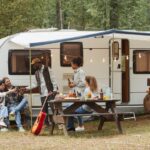 A diverse group of young people camping by an RV in a forest, relaxing outdoors with plenty of open copy space.