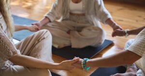 A group of people in leisurely clothing sitting on a wooden floor and holding hands to form a circle.