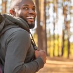 A smiling college-age person looking over their shoulder as they hike through a wooded area while wearing a backpack.